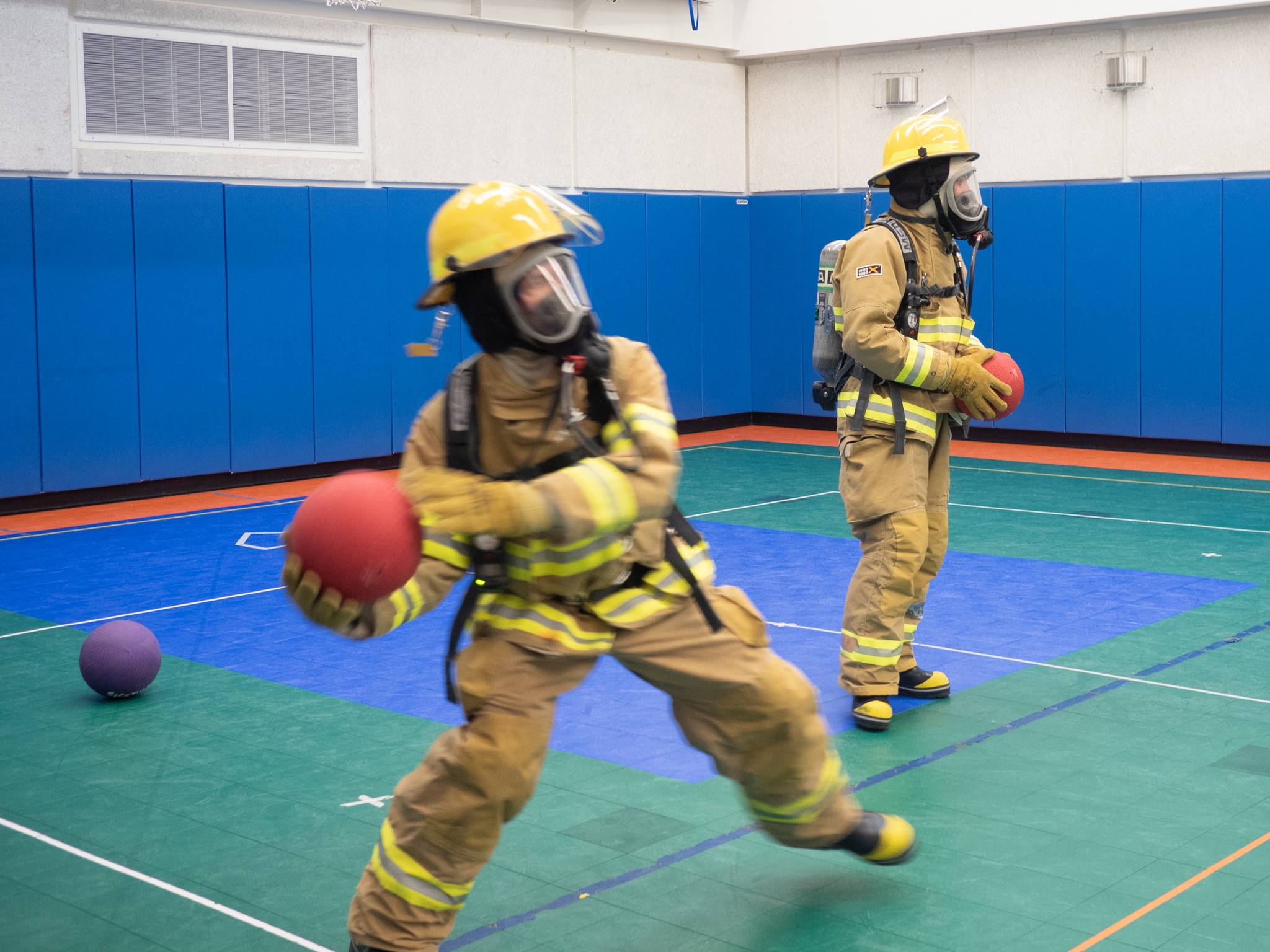 Two people in fire gear playing dodgeball in gym.]