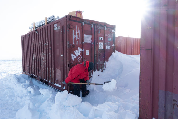 Person in red parka shoveling snow outside the doors of a shipping container.