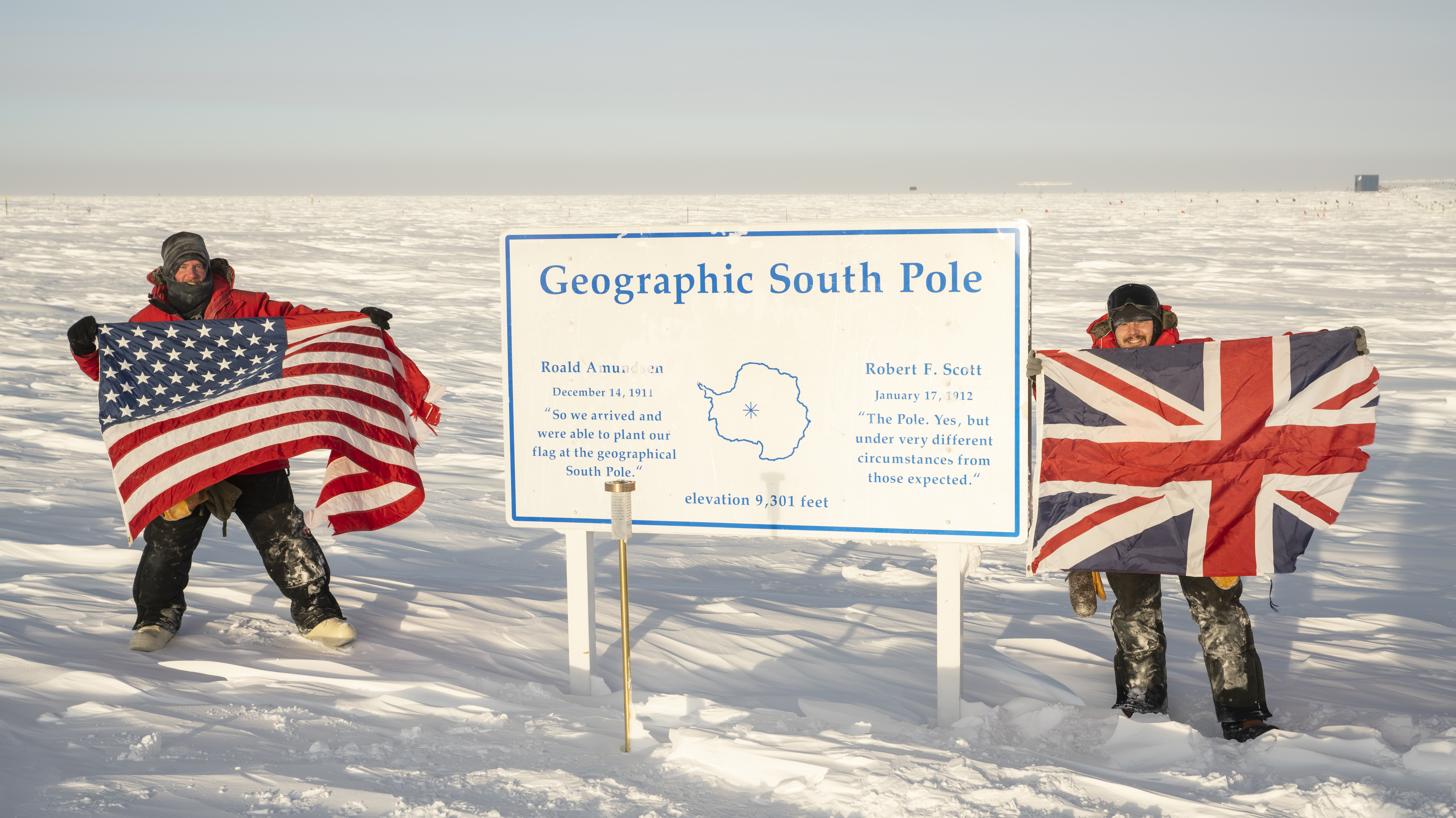 IceCube winterovers holding US and British flags next to geographic South Pole sign.