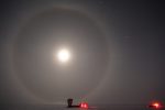 A bright moon with large moon halo at the South Pole, with South Pole Telescope seen in shadow in the distance.
